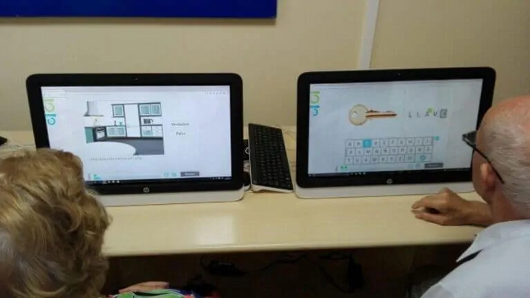 Due computer su tavolo educativo; gli schermi mostrano un progetto di cucina e un tasto, con due utenti di fronte a essi.