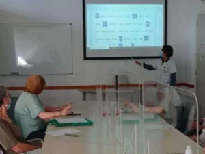 Gruppo di partecipanti in una sala riunioni osservando una presentazione; una persona in piedi indica lo schermo, con mascherine e paraventi.