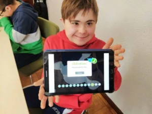 Bambino sorridente con felpa rossa tiene una tablet in un'aula; fa parte di un gruppo di persone con sindrome di Down, ambiente di apprendimento.