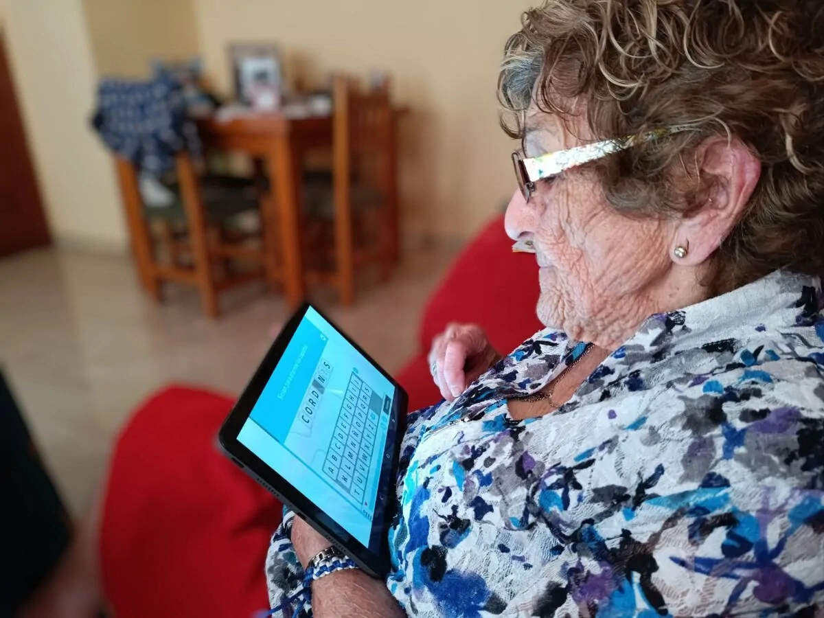 Donna anziana seduta su divano rosso usa tablet, occhiali e capelli ricci, concentrata sullo schermo; sfondo con tavolo e sedie.