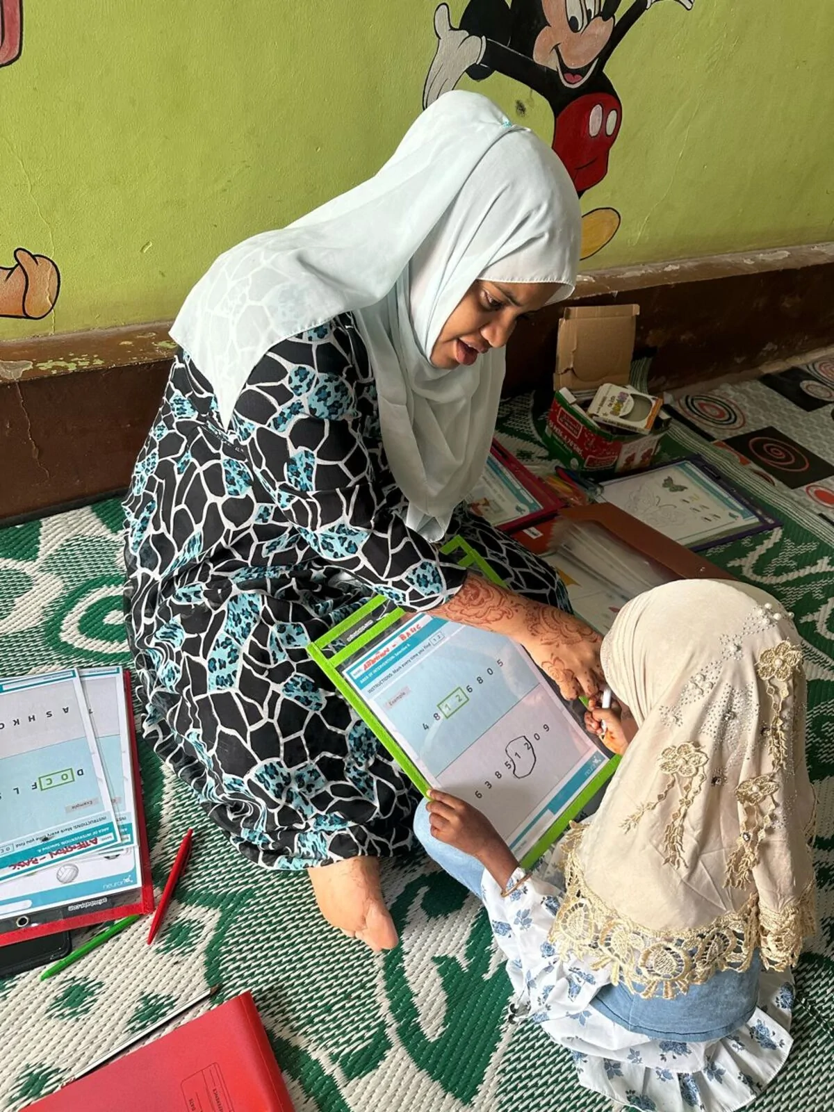 Donna con hijab seduta su un tappeto insegna una bambina, con materiali didattici e decorazioni murali sullo sfondo.