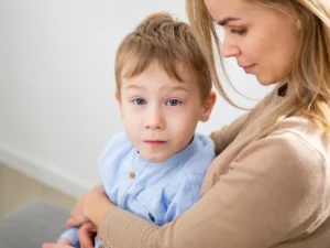 Illustrazione educativa: donna che abbraccia un bambino in un ambiente domestico, sfondo chiaro ed espressione neutra.