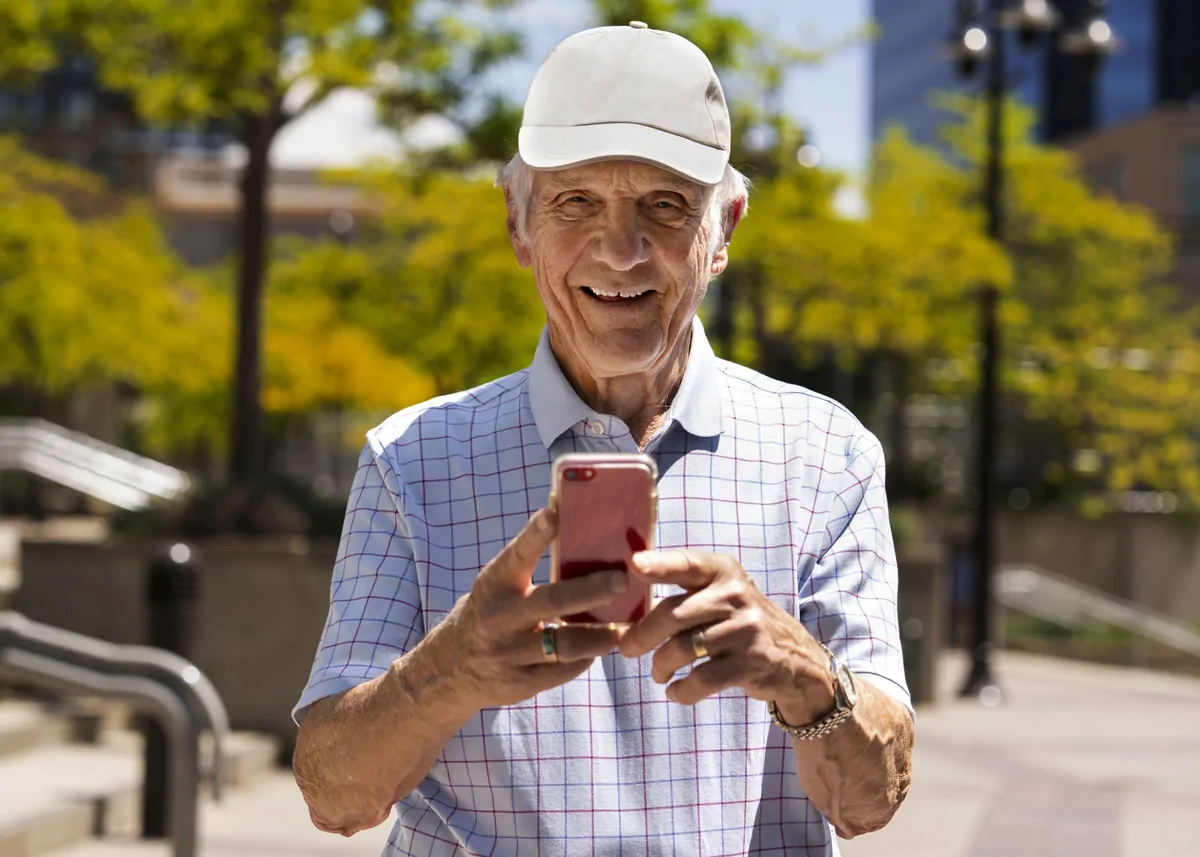 Uomo anziano sorridente che tiene un telefono cellulare in primo piano, camicia a quadri e cappellino bianco, sfondo urbano soleggiato.