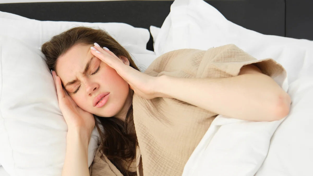 Donna giovane a letto, mani sulle tempie, espressione di disagio, avvolta in coperta, cuscini bianchi.