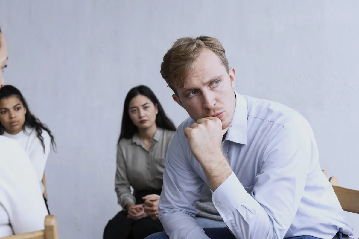Gruppo di tre persone in un ambiente interno: uomo in primo piano con mento appoggiato, due donne dietro lo osservano, atmosfera riflessiva.