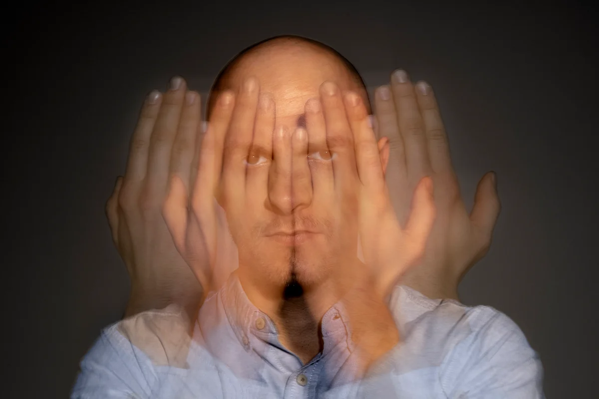 Uomo adulto con testa rasata guarda l'obiettivo, mani sfocate sovrapposte per movimento, sfondo scuro che evidenzia il soggetto.
