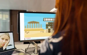 Persona al tavolo davanti al computer: puzzle di lettere sullo schermo e paesaggio con tempio; laptop mostra il volto di una giovane.