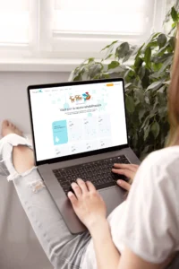 Persona seduta a gambe incrociate davanti a laptop argentato, aperto su pagina neuroriabilitazione, con finestra luminosa e piante.