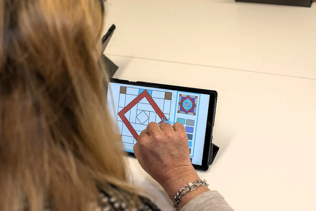 Persona che usa tablet su tavolo in ambiente di lavoro; schermo mostra disegno colorato con forme geometriche.