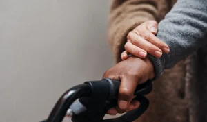 Due mani intrecciate in primo piano, una delle quali sostiene un oggetto; tono della pelle chiaro e scuro, sfondo neutro.