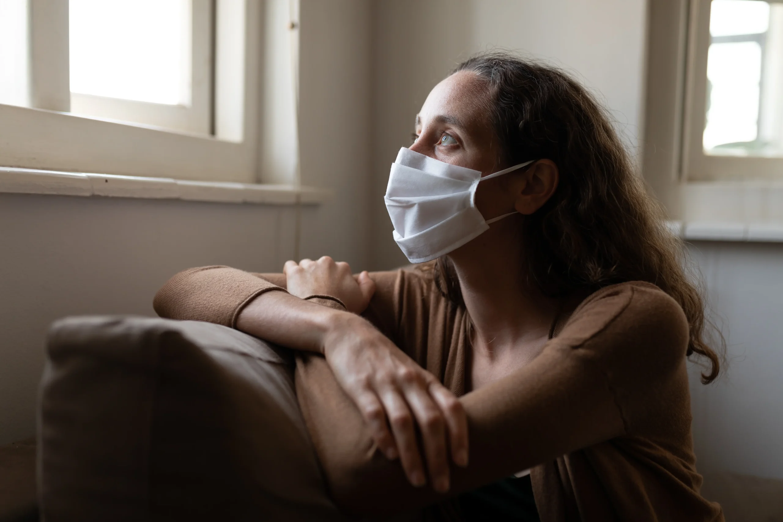 Donna con una mascherina bianca seduta su un divano, che guarda verso la finestra con un'illuminazione soffusa in un ambiente interno semplice.