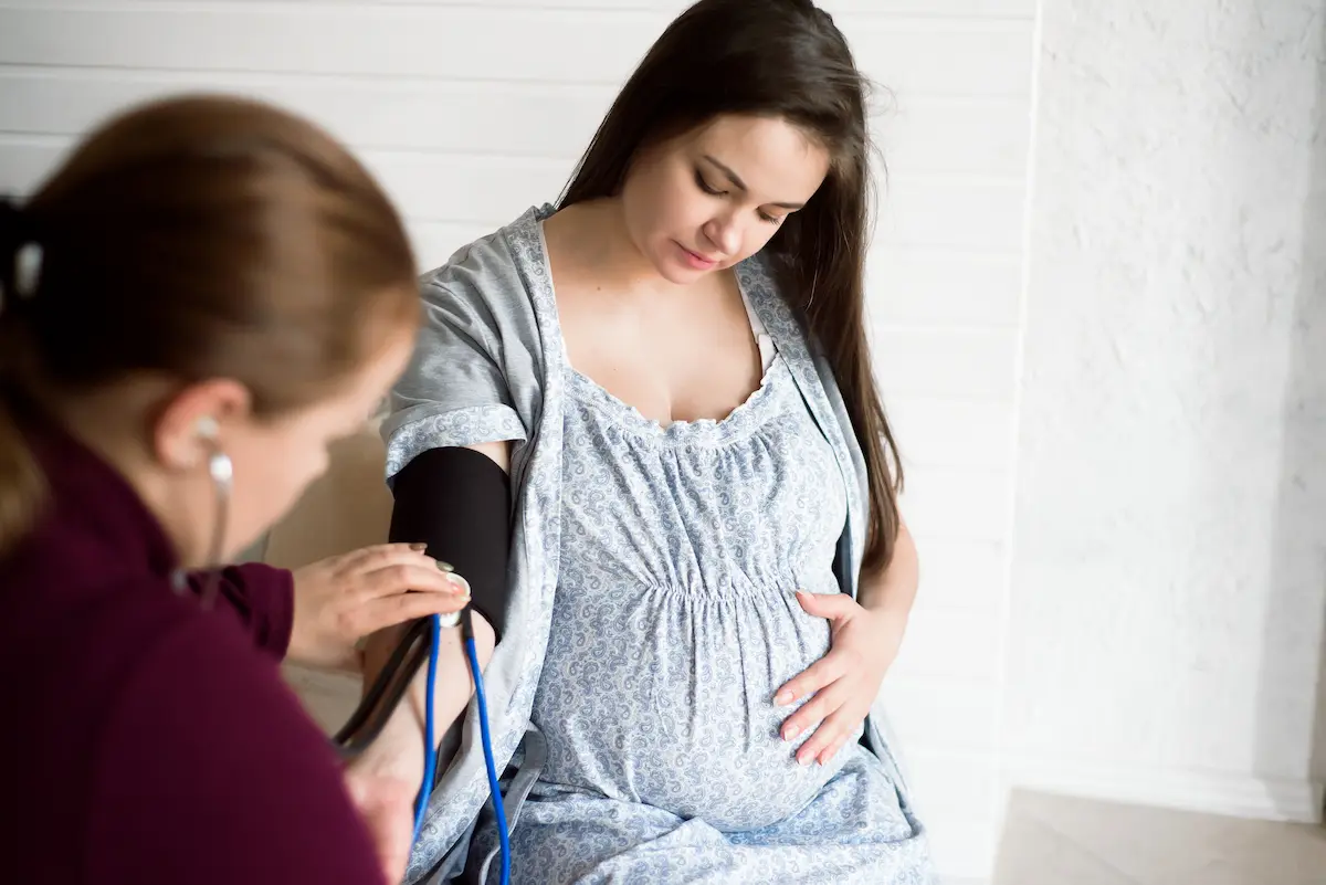 Una donna incinta riceve assistenza medica mentre un'operatrice sanitaria le rileva la pressione sanguigna; a sinistra, un flacone di colina.