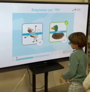 Bambino interagisce davanti a uno schermo durante una sessione di NeuronUP; si vede una scimmia e un'anatra, con un esercizio di parole che iniziano con PA.