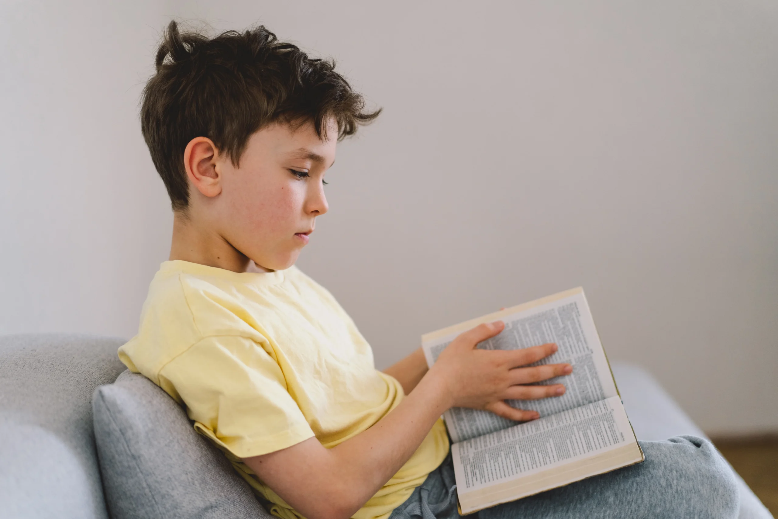 Ragazzo seduto su un divano, maglietta gialla e capelli castani disordinati, che legge un libro; ambiente moderno e tranquillo.