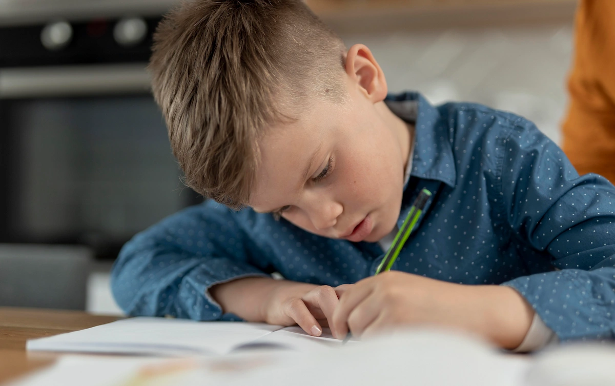 Bambino concentrato a scrivere o disegnare su un foglio, camicia azzurra con puntini, in un ambiente domestico accanto a icone di apprendimento e benessere mentale.