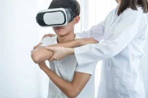 Молодой человек в VR-шлеме, сосредоточенно выполняющий упражнение; специалист поддерживает его руки во время реабилитации.