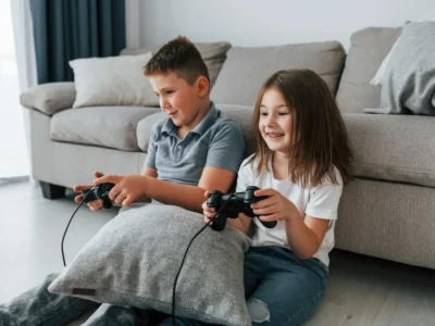 Двое детей сидят на полу и играют с игровыми контроллерами перед серым диваном и подушками при мягком освещении.