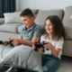 Двое детей сидят на полу и играют с игровыми контроллерами перед серым диваном и подушками при мягком освещении.