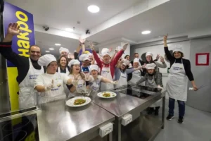 Группа людей с синдромом Дауна на кухне в фартуках и поварских колпаках празднует за рабочим столом.