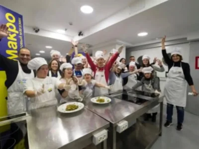 Группа людей с синдромом Дауна участвует в кулинарном занятии в белых поварских колпаках и фартуках в радостной атмосфере.