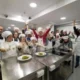 Группа людей с синдромом Дауна участвует в кулинарном занятии в белых поварских колпаках и фартуках в радостной атмосфере.