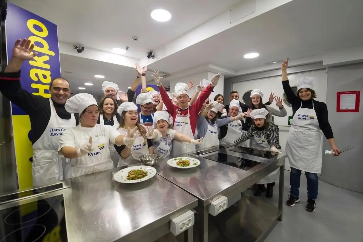 Группа людей с синдромом Дауна на кухне в фартуках и поварских колпаках празднует за рабочим столом.