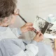 Mujer mayor sentada mirando una foto en blanco y negro; lleva gafas, cabello canoso y bastón; mesa con más fotos y una taza.