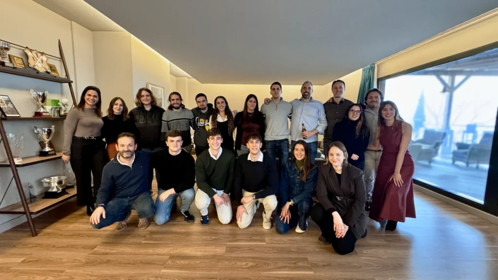 Grupo grande de personas posando en interior con luz natural, organizadas en dos filas (de pie y sentadas), fondo con estanterías y trofeos.