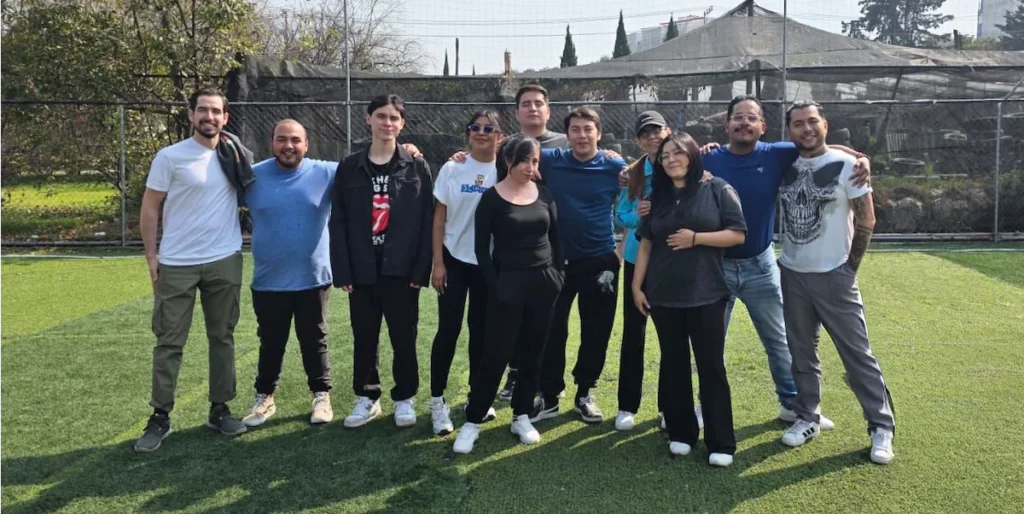 Grupo de personas posando al aire libre sobre césped, ropa casual, con varias sonrisas; fondo con red deportiva y árboles.