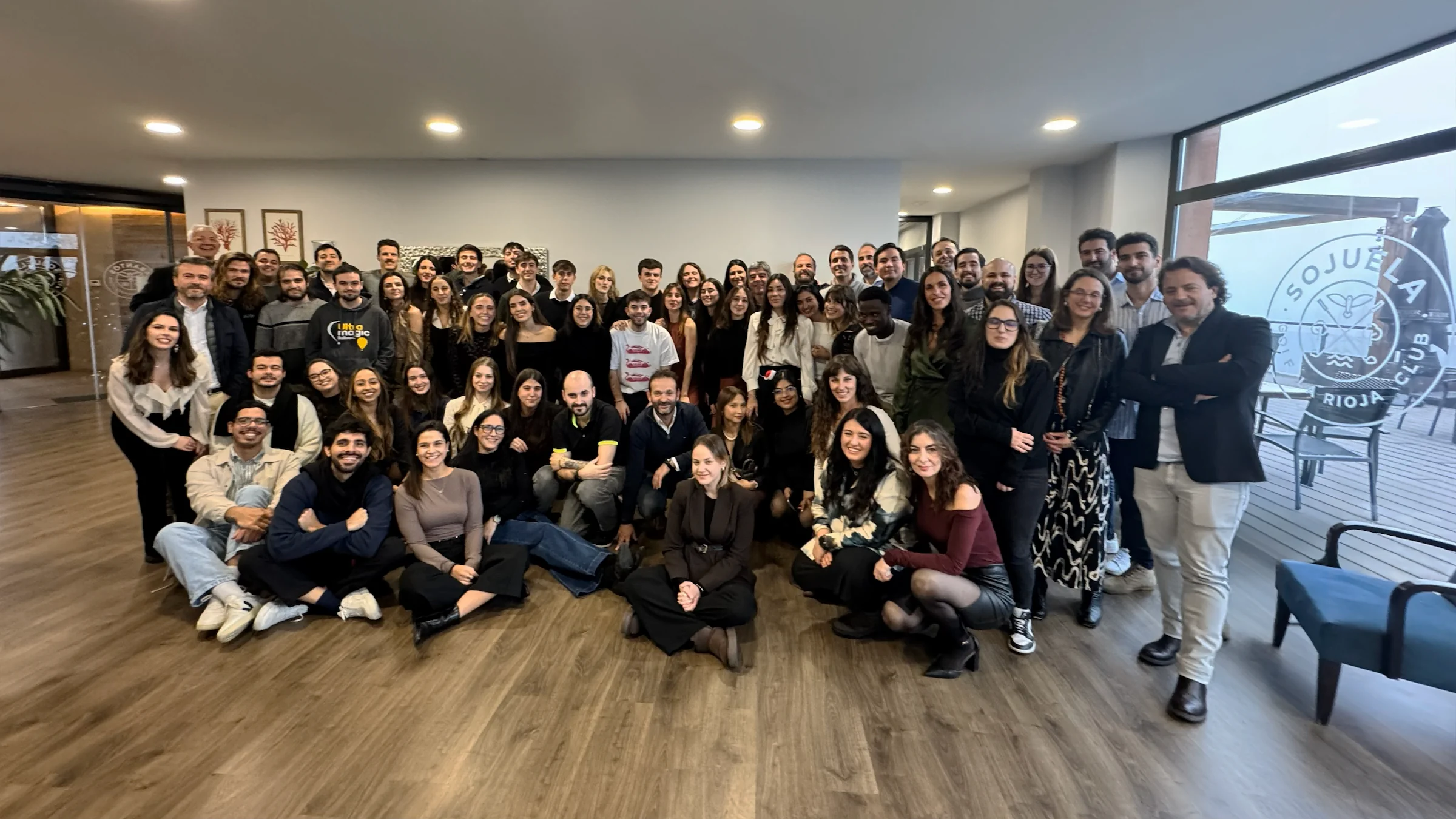 Grupo grande posando en interior moderno con suelo de madera; personas vestidas casual, sonriendo, con plantas al fondo.